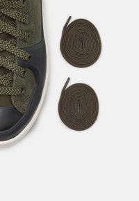 Baskets vertes avec tige en tissu texturé, bout en caoutchouc noir, et lacets olive, accompagnées de deux rouleaux de lacets assortis.