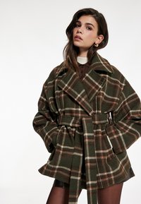 Abrigo de lana a cuadros en verde oscuro con detalles en beige y marrón. Cuenta con solapas anchas, cintura con cinturón y grandes bolsillos laterales para mayor practicidad.