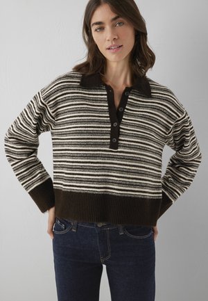 Young woman wearing a striped brown and beige knit sweater with buttons and dark blue jeans standing against a plain wall.