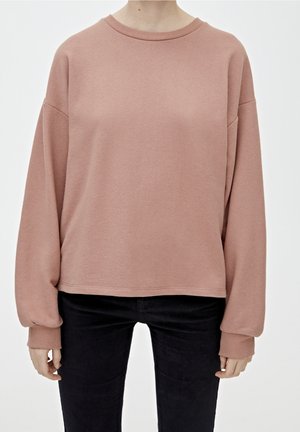 Personne portant un sweat-shirt ample à manches longues rose poussiéreux avec des poignets côtelés, associé à un pantalon noir ajusté, debout devant un fond uni.