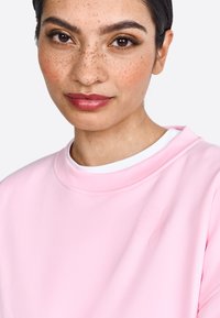 Felpa rosa chiaro con una texture morbida, caratterizzata da un design floreale in rilievo sulla sinistra. Collo a giro e sottocamicia bianca a strati visibile.
