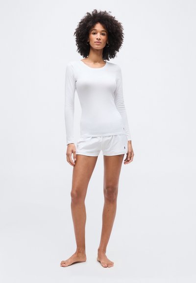 Γυναίκα με σγουρά μαλλιά, όρθια και ξυπόλητη, φοράει λευκό πουκάμισο με μακριά μανίκια και λευκές σορτς μπροστά σε απλό φόντο.