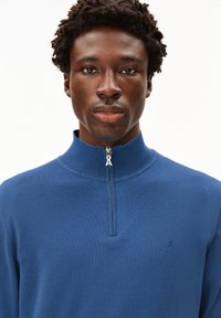 Maglione blu con zip e collo alto, realizzato in morbido materiale lavorato a maglia. Presenta un piccolo logo sul lato sinistro e un dettaglio del tirazip.