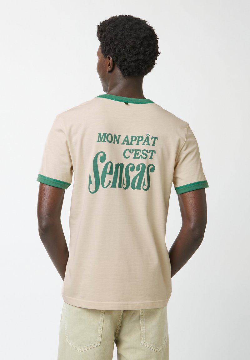 Beige katoenen t-shirt met groene ribbels aan de kraag en mouwen. De achterkant heeft bedrukte tekst: "MON APPÂT C'EST Sensas".