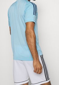 Camiseta atlética de color azul claro con textura de malla, de mangas cortas y rayas azul marino en los hombros. Combinada con pantalones cortos blancos que tienen acentos en negro.