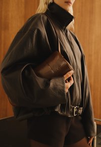 Γυναίκα ντυμένη με υπερμεγέθη μαύρο δερμάτινο μπουφάν και μαύρα σορτσάκια κρατώντας μια μικρή καφέ δερμάτινη τσάντα μπροστά σε ξύλινο φόντο.