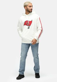 Weißer Hoodie mit rotem Grafikdesign der Buccaneers-Flagge, ausgestattet mit roten Seitestreifen, einer vorderen Tasche und gerippten Bündchen, kombiniert mit hellblauen Jeans und schwarzen Schuhen.
