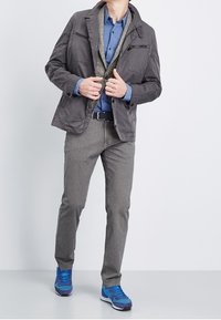 Giacca grigia con tasche zip sopra una camicia blu a bottoni, pantaloni grigi e scarpe da ginnastica blu con dettagli viola. Dettagli testurizzati visibili.