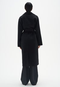 Manteau noir ceinturé avec une silhouette structurée, des manches longues et un ourlet droit, fabriqué à partir d'un tissu texturé, vu de dos.
