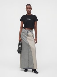 Woman stands wearing black cropped t-shirt, two-tone long denim skirt, black ankle boots, and holding small black handbag.