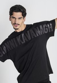 Camiseta de gran tamaño negra hecha de algodón, con un gran logo en relieve "GIANNI KAVANAGH" en el pecho. Corte holgado con costuras de hombro caídas.