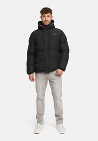 Mand står iført en sort polstret jakke, lys grå jeans og hvide sneakers foran en ensfarvet hvid baggrund.