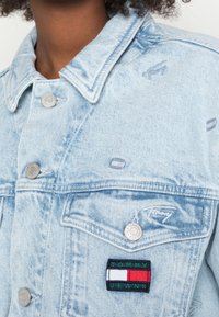 Ljusblå denimjacka med knäppning och Tommy Jeans-märke på vänster knappficka, bärs av person med lockigt hår.