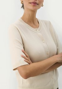 Cardigan beige à manches courtes, avec bordure côtelée et cinq boutons. Texture en tricot lisse et léger, avec un col rond.
