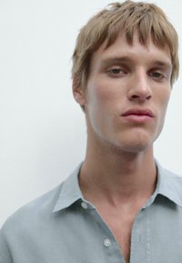 Homme avec des cheveux châtains clairs courts et ébouriffés, portant une chemise à boutons bleu pâle. Le tissu semble doux et légèrement froissé. Fond neutre.