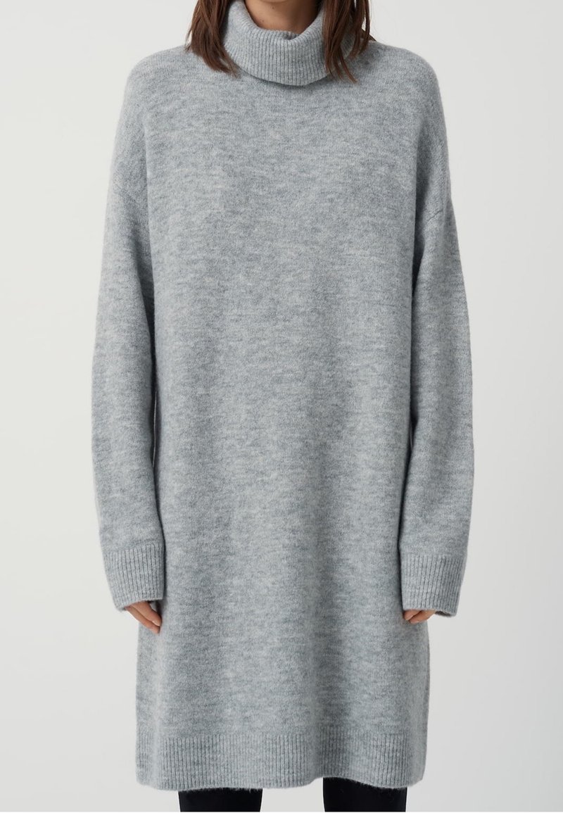 Personne portant une longue robe-pull à col roulé tricotée gris clair avec des poignets et un ourlet côtelés, debout devant un fond uni.