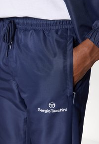 Tamsiai mėlynos sportinės kelnės, pagamintos iš tekstūruoto audinio, su elastingu juosmeniu, šonine kišene ir baltu "Sergio Tacchini" logotipo akcentu.