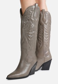 Bottes de cowboy en cuir gris avec des coutures surélevées, un bout pointu et un talon incliné. Présente des découpes décoratives et une texture lisse.
