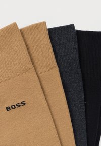 Trzy pary prążkowanych skarpet w kolorach beżowym, ciemnoszarym i czarnym, z małym czarnym logo na beżowej parze. Każda skarpeta ma dzianą teksturę.