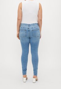 Kvinna i ljusblå tajta jeans, vit ärmlös topp och vita klacksandaler, sedd bakifrån mot en neutral bakgrund.