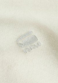 Close-up of light beige fabric with a stitched abstract logo and the word "PRYM" embroidered in matching thread.