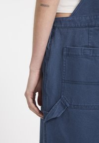 Denim schürze in donkerblauw met een zijzak en stiksels. De stof heeft een gestructureerde afwerking en is lichtgewicht.