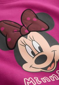 Cara de Minnie Mouse con un gran lazo rojo con lunares rosas sobre un fondo púrpura, sonriendo con los ojos mirando de lado.