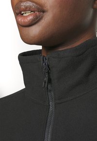 Schwarze Fleece-Jacke mit hohem Kragen, ausgestattet mit einem Frontreißverschluss, glatter Textur und strukturiertem Ausschnitt. Der Fokus liegt auf funktionalen Details.