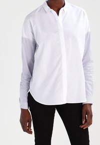 Bílá košile s dlouhými pruhovanými rukávy, s límečkem na knoflíky a zaobleným lemem. Hladká bavlněná textura s kontrastním designem.