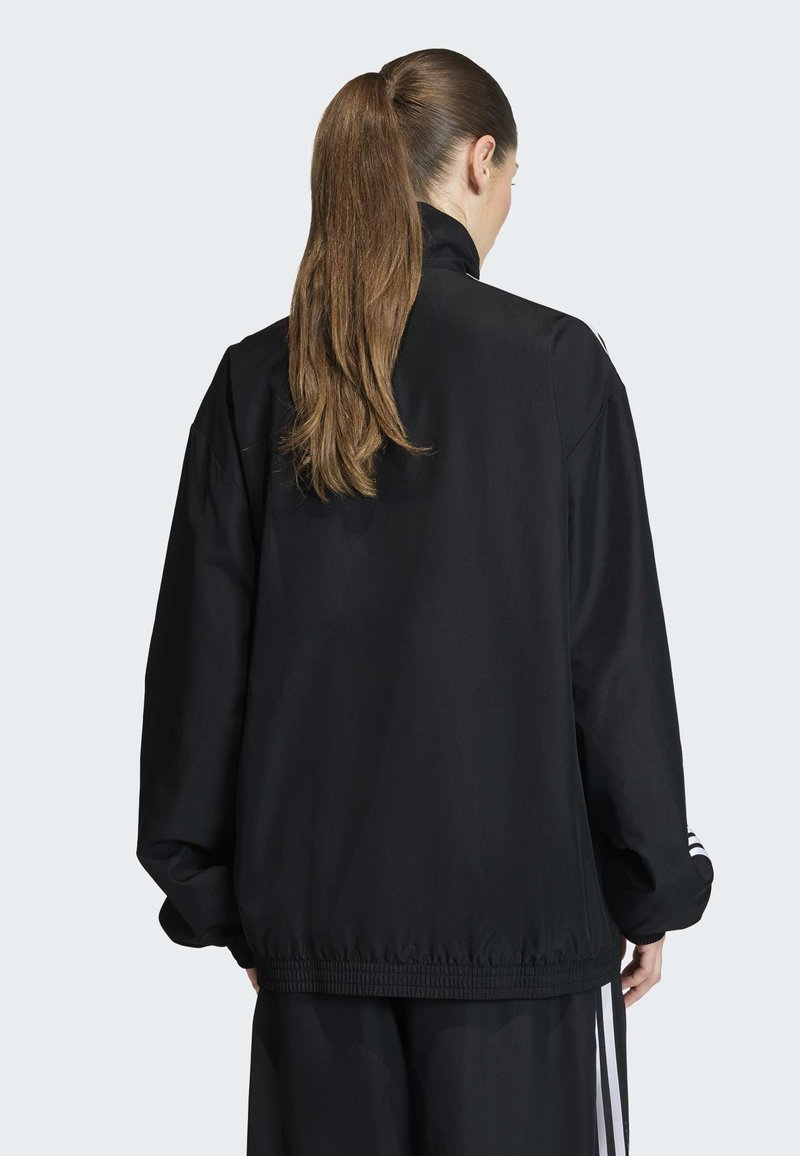 Persona con una lunga coda di cavallo marrone che indossa una giacca sportiva nera e pantaloni neri abbinati con strisce bianche, vista da dietro.