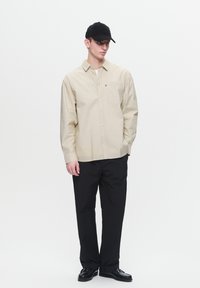 Beige overhemd met lange mouwen en subtiele strepen, enkele zak; gecombineerd met zwarte broek en zwarte pet. Zwarte schoenen maken de look compleet.
