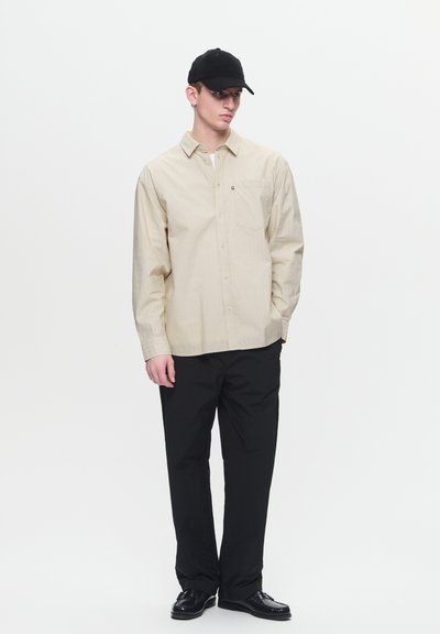 Chemise beige à manches longues avec des rayures discrètes, poche unique ; associée à un pantalon noir et une casquette noire. Des chaussures noires complètent le look.