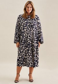 Robe midi bleu marine avec un motif abstrait crème, manches longues, devant boutonné et coupe ample, associée à des talons noirs à bride cheville.