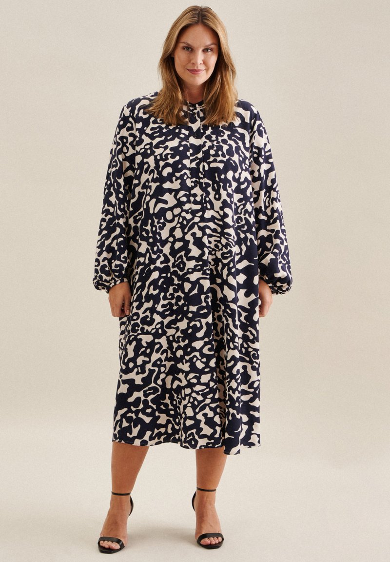 Robe midi bleu marine avec un motif abstrait crème, manches longues, devant boutonné et coupe ample, associée à des talons noirs à bride cheville.