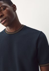 Maglietta di colore blu scuro con tessuto strutturato, collo a girocostine con righe dorate e marroni e piccolo logo in rilievo sul petto.