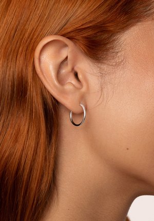 Gros plan sur l'oreille d'une personne aux cheveux roux, portant une boucle d'oreille simple en argent en forme d'anneau sur le lobe de l'oreille.