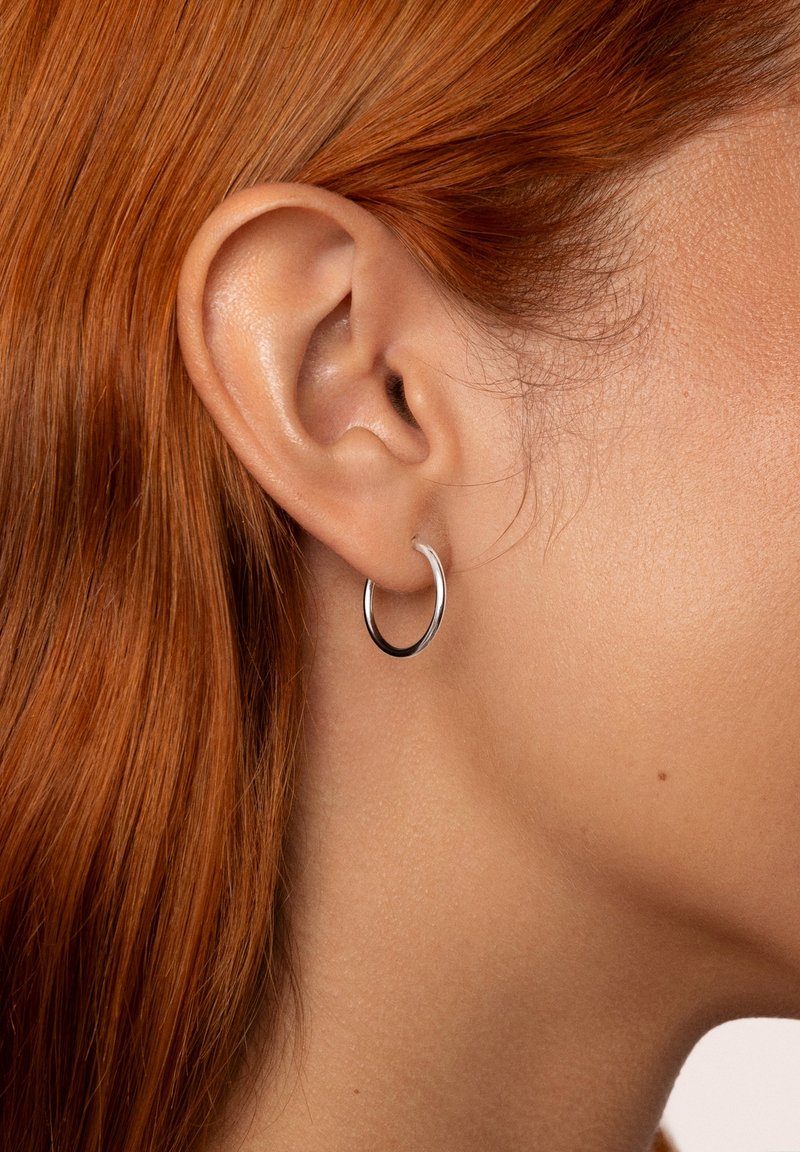 Gros plan sur l'oreille d'une personne aux cheveux roux, portant une boucle d'oreille simple en argent en forme d'anneau sur le lobe de l'oreille.