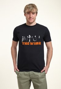 Svart bomulls-t-shirt med en grafisk bild av karaktärer från "The Wire" i vit och orange text på framsidan. Kortsleeves, rundad halsdesign.
