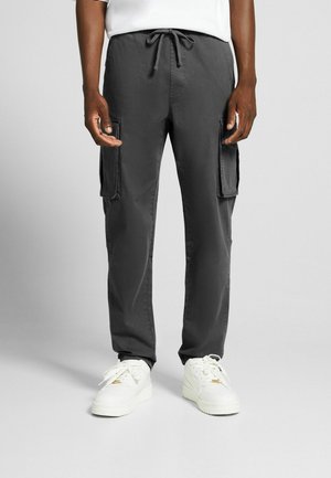 Gray cargo pants with a drawstring waist, featuring two large side pockets and tapered legs; worn with white sneakers. Soft fabric texture.