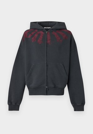 Personne portant un sweat à capuche noir oversize zippé, un jean gris foncé ample, des baskets rouges et de grandes lunettes de soleil noires, debout devant un fond uni.