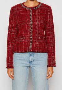 Vrouw in een gestructureerde rode tweedjack met kettingafwerking, gecombineerd met lichtblauwe high-waisted spijkerbroek, staand tegen een effen achtergrond.