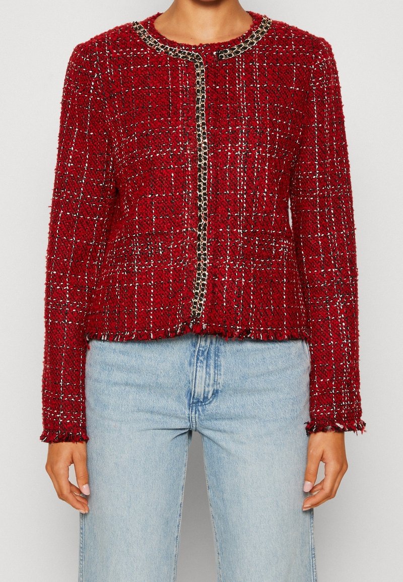 Vrouw in een gestructureerde rode tweedjack met kettingafwerking, gecombineerd met lichtblauwe high-waisted spijkerbroek, staand tegen een effen achtergrond.