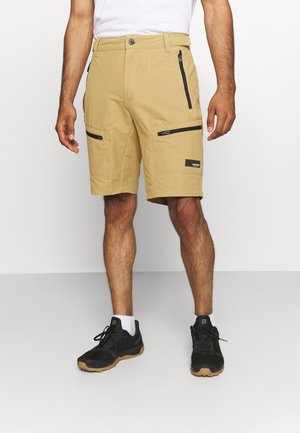 Hombre vestido con pantalones cortos color canela hasta la rodilla con bolsillos con cremallera negros, calcetines blancos y zapatos deportivos negros, de pie contra un fondo liso.