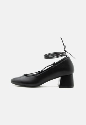 Schwarzer Lederschuh mit mittlerem Absatz, spitzer Schuhspitze, gekreuzten Riemen und Wickelverschluss um den Knöchel auf weißem Hintergrund.