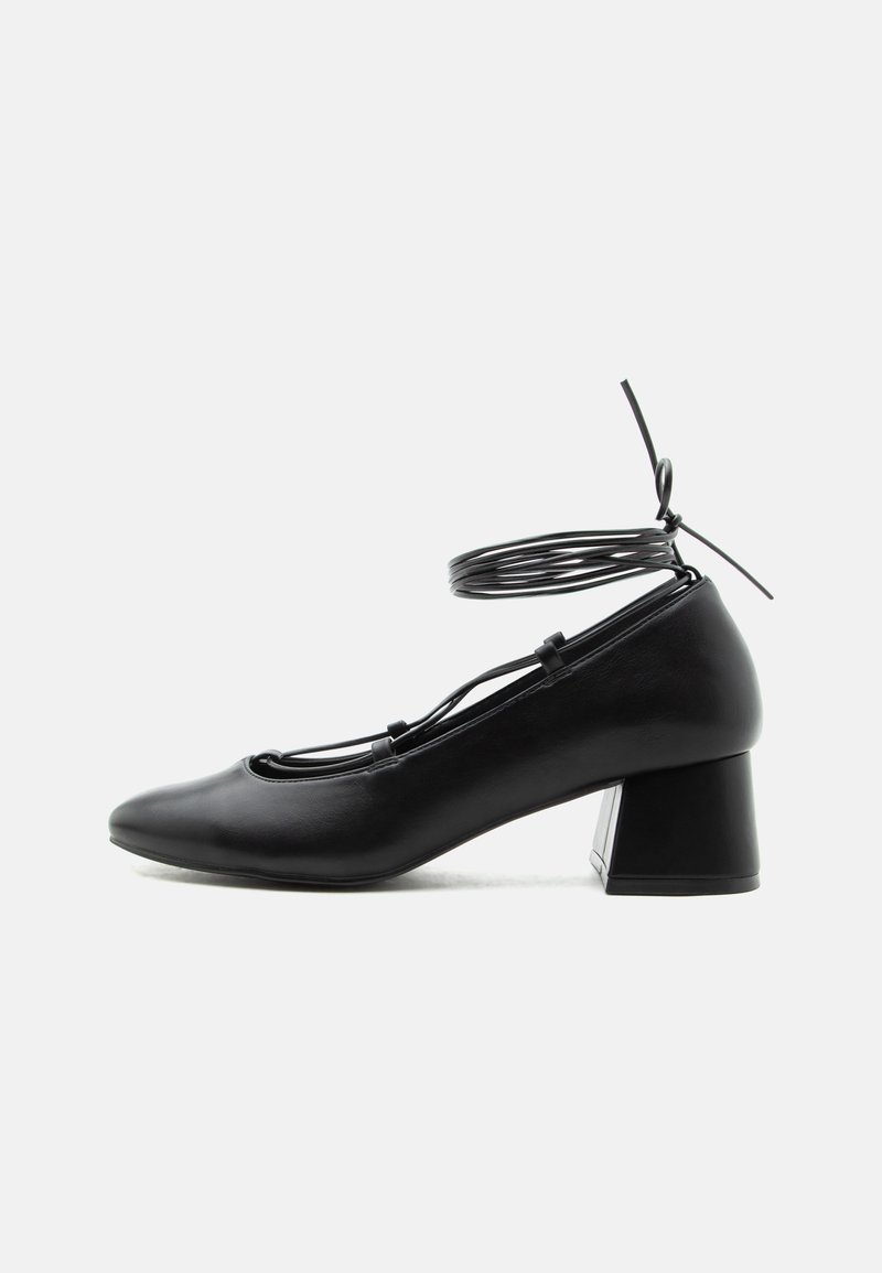 Μαύρο δερμάτινο παπούτσι με μεσαίο τακούνι, μυτερή μύτη, σταυρωτά λουριά και δεσίματα γύρω από τον αστράγαλο σε λευκό φόντο.