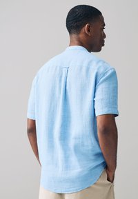 Kortærmet skjorte i lyseblåt stof med en tekstureret vævning. Har en rygskulder og en buet sømkant, kombineret med beige bukser.