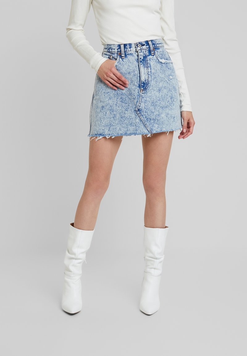 Minigonna in denim lavaggio chiaro con orlo sfrangiato, vita alta e tasche anteriori, abbinata a stivali bianchi al ginocchio.