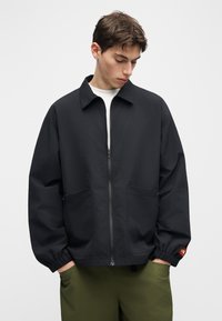 Νεαρός άνδρας κοιτάζει προς τα κάτω φορώντας μαύρο μπουφάν με φερμουάρ και ελαστικές μανσέτες, καθώς και πράσινο παντελόνι, με τα χέρια στις τσέπες.