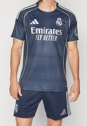 Mann trägt dunkelblaues Real Madrid Fußballtrikot und passende Shorts mit Team- und Adidas-Logos, steht vor einem einfarbigen Hintergrund.