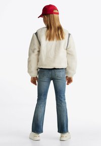 Biała polarowa kurtka w czarne paski, niebieskie dżinsy dzwony, białe buty i czerwona czapka. Widok z tyłu ukazuje długie włosy i luźny krój.